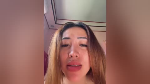 A video of an Asian woman with long, straight, light brown hair, fair skin, and a round face. She has a nose ring and is smiling slightly. The background is a blurred indoor setting with a ceiling light.