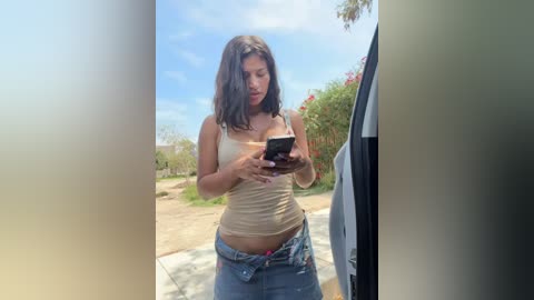 Video of a young woman with medium-length brown hair, wearing a beige tank top and blue jeans, checking her smartphone while standing outside a car in a suburban area.