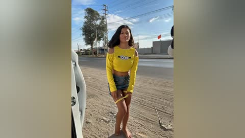 Video of a Latina woman with long black hair, wearing a yellow off-shoulder crop top and denim shorts, standing on a dusty road with utility poles and trees in the background.