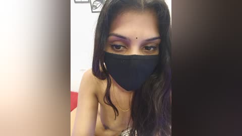 Media: Video of a topless woman with long, dark hair, wearing a black face mask and bold eye makeup, standing in a dimly lit room.