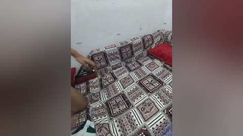 Video of a person, possibly female, with light skin tone, lying on a red and white patterned quilted bedspread in a dimly lit room with a red cushion and a white wall.