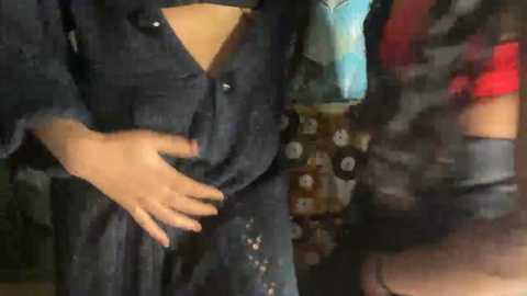 Video of a person in a dark robe with hands on their belly, standing in a cluttered room with colorful, patterned objects and a red shirt partially visible.