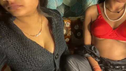 Video of two women seated closely; one in a dark knit cardigan, the other in a red bra, both with pearl necklaces.