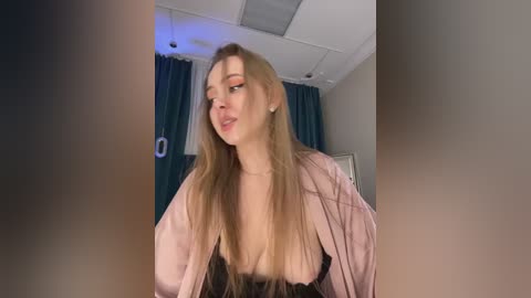 A video of a fair-skinned, blonde woman with long hair, wearing a pink robe, black lingerie, and makeup, standing in a room with teal curtains and a ceiling light.