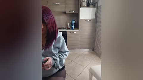 Video of a woman with red hair, wearing a grey hoodie, sitting on a white chair in a modern kitchen, holding a green bottle.