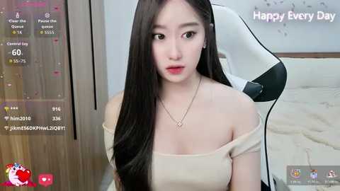 A video of an East Asian woman with long black hair, fair skin, and a slender physique, wearing a beige off-shoulder top, sitting on a white gaming chair in a minimalist bedroom.