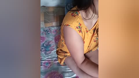 Video of a young woman with light skin, wearing a yellow floral blouse, sitting on a bed with a colorful, patterned sheet.