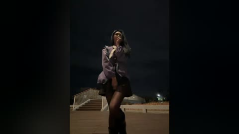 A dark video of a woman in a purple coat and short skirt, standing on a dimly lit industrial site with metal structures and stairs in the background.
