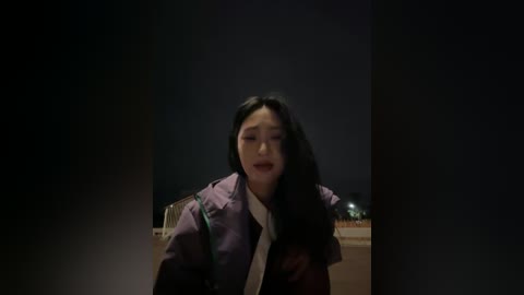 Video of an Asian woman with long black hair, wearing a purple jacket and white shirt, standing against a dark, night sky background with faint city lights visible.