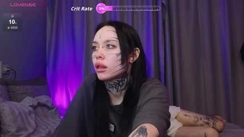 Video of a pale-skinned, black-haired woman with a tattooed neck, wearing glasses, in a dimly-lit room with purple lighting, holding a gray pillow, on a bed with gray sheets.
