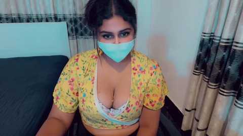 Video of a plus-sized woman with dark skin and long black hair, wearing a floral crop top, revealing large breasts, and a blue face mask. Background shows a bed with dark sheets and beige curtains.