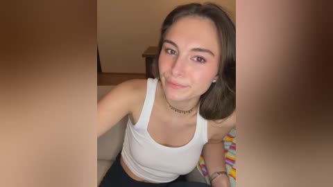 A video of a young woman with light skin, shoulder-length brown hair, and a white tank top, sitting indoors, smiling slightly.