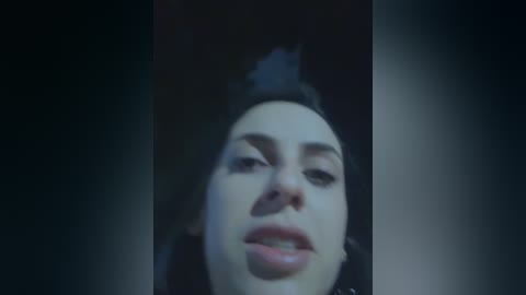 Video of a close-up face with dark hair, fair skin, and full lips, set against a dark, indistinct background. The image is dimly lit, creating a moody atmosphere.