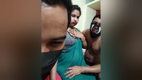Video: A distressed woman in a teal sari, wearing a gray bra, is held by two men in a room. One man is shirtless, wearing a black mask, and the other has a dark face mask.
