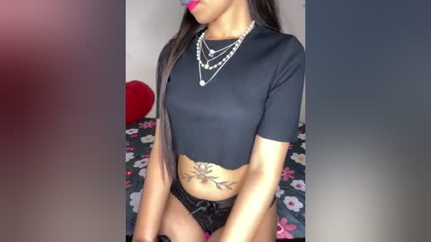 Video of a light-skinned woman with long, straight black hair, wearing a black crop top, black lace panties, and multiple necklaces. She sits on a bed with a floral-patterned duvet.