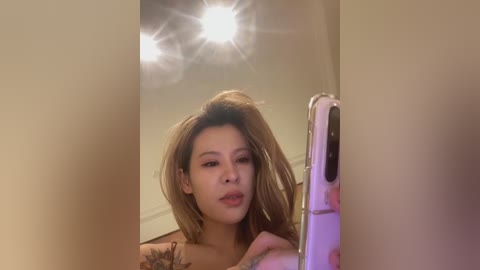 Video of a young Asian woman with light brown hair, styled in a voluminous, messy top knot. She has a tattoo on her left arm, and is taking a selfie in a dimly lit room with a ceiling light creating a starburst effect.