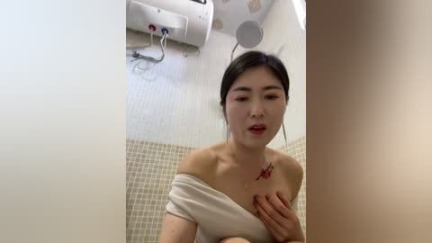 A video of an Asian woman with dark hair, wearing a white off-shoulder top, sitting on a beige-tiled floor, with a red mark on her chest, and a white wall-mounted heater in the background.
