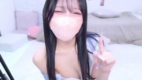 A video of an Asian woman with long black hair, wearing a white top, blowing a bubble, in a bedroom with a bed and white walls.