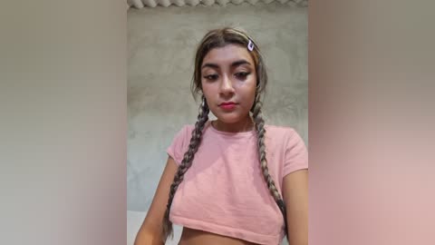 Video of a young Latina woman with light brown skin and long, braided hair. She wears a light pink crop top, standing in front of a textured, off-white wall.