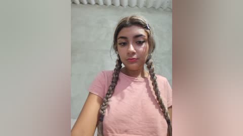 Video of a young woman with light brown skin, wearing twin braids, a pink t-shirt, and standing against a light beige wall with a tiled ceiling.