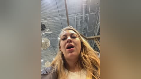 Video of a blonde woman with long hair, wearing a gray jacket, looking surprised and mouth open, indoors with a high ceiling, exposed pipes, and a chandelier visible in the background.