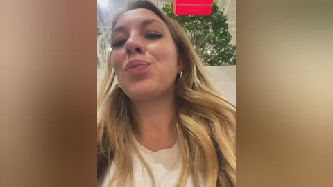 Video of a blonde woman with light skin, wearing a white shirt, making a pouty face. Background features indoor plants and a red sign.