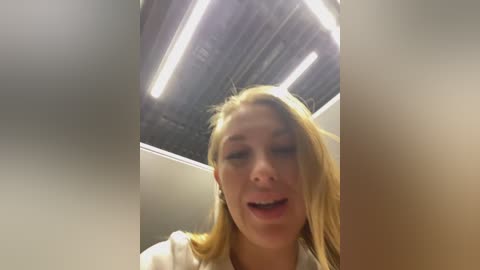 Video of a young Caucasian woman with long blonde hair, wearing a white shirt, smiling, captured from a low angle, with fluorescent lighting and a tiled ceiling in the background.
