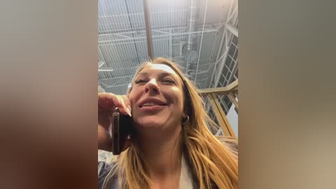 Video of a smiling woman with long blonde hair and light skin, holding a phone to her ear in a modern, well-lit room with metal grids on the ceiling.