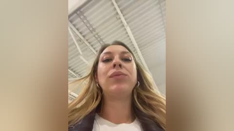 Video of a young woman with long, wavy blonde hair, fair skin, and light makeup, wearing a white top and a dark jacket. She gazes upwards with a neutral expression. Background shows a white ceiling with visible beams.