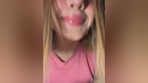 A close-up video of a young woman with long, blonde hair and fair skin, wearing a pink shirt, smiling widely with her mouth open. The background is blurred and brown.
