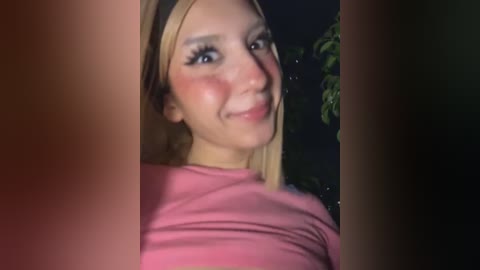 Video of a smiling young woman with fair skin, rosy cheeks, and long blonde hair, wearing a pink top. Background is dark, with blurred green foliage.