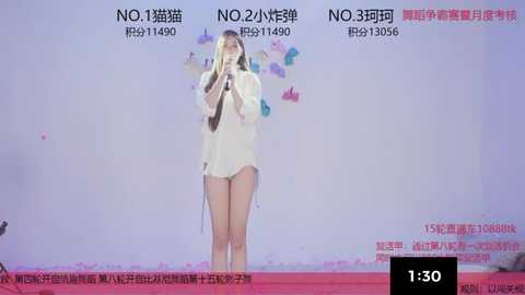 A video of an Asian woman singing into a microphone on stage, wearing a white dress, surrounded by pink and purple balloons. The background is a soft pastel pink, with text in Chinese.
