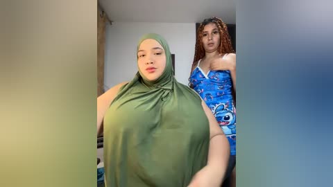Video of a plus-sized woman with fair skin and long curly hair, wearing a green hijab, standing beside a curvy woman with medium skin tone and dreadlocks in a blue patterned dress.