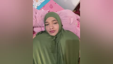 Video of a young woman with medium skin tone and a round face, wearing a green hijab, lying on a pink bed with floral-patterned sheets.