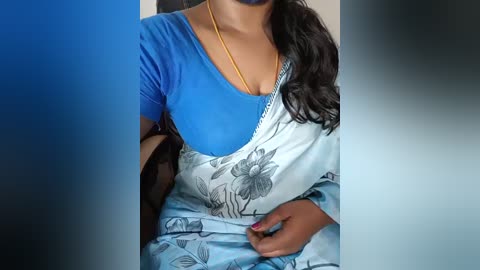 Video of a woman with medium brown skin, wearing a blue top and white sari with floral print, gold necklace, and purple nail polish.