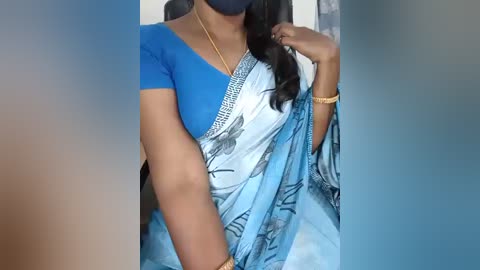 Video of a South Asian woman wearing a vibrant blue sari with intricate black patterns, a blue short-sleeve blouse, gold bracelets, and a dark blue face mask. She has medium brown skin and long, wavy black hair.