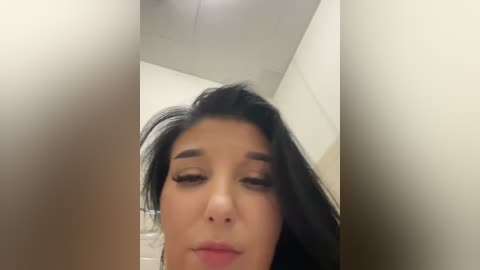 A close-up video of a young woman with long, dark hair and light skin, her eyes closed and lips slightly parted, standing in a bathroom with a beige tiled wall and white ceiling.