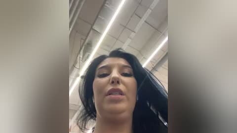 A video captures a low-angle view of a woman with long, black hair and full lips, wearing a light-colored top, standing in a modern, industrial-style room with exposed pipes and fluorescent lighting.