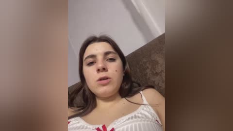 Video of a young woman with fair skin, dark hair, wearing a white bra with red accents, lying on a brown carpet. Background shows a white ceiling and beige wall.