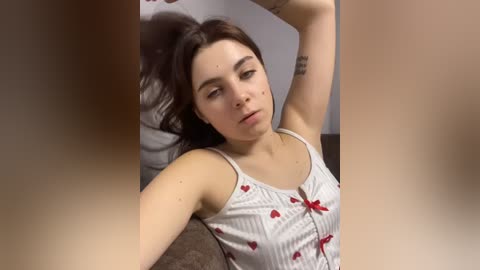 Video of a fair-skinned, dark-haired woman with a tattoo on her left arm, wearing a white tank top with red heart patterns, lying on a bed, looking up.