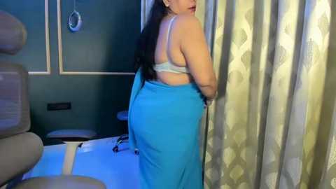 Video of a woman in a medical setting. She has medium skin tone, long dark hair, and is wearing a light blue towel wrapped around her waist, partially revealing a white bra.