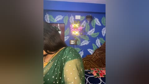 Video of a woman with dark hair, wearing a green sari with white polka dots, standing in a room with blue walls adorned with leafy decals and a \"Stay Strong\" neon sign.