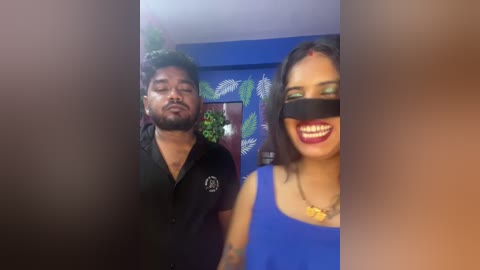 Video of a man in a black polo shirt and woman in a blue tank top, both wearing black masks, standing in a room with blue walls and fern paintings.