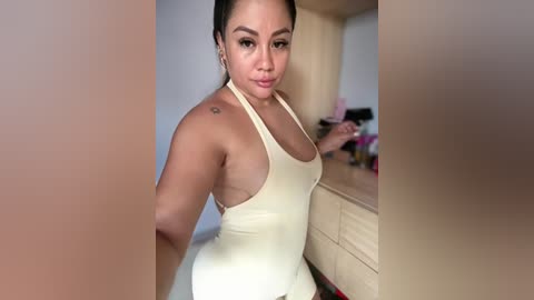 Video of a light-skinned woman with medium-length black hair in a white halter top, standing indoors near a wooden cabinet.