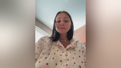 Video of a young woman with medium skin tone, straight black hair, wearing a white button-up shirt with small red hearts. Background shows a blurred indoor setting with soft lighting.