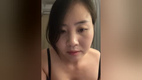 Video of a young East Asian woman with straight black hair, wearing a black spaghetti strap top, looking down with a neutral expression in a dimly lit room.