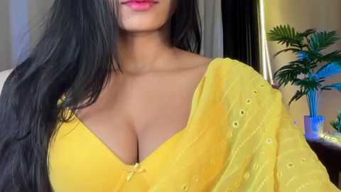 Video of a South Asian woman with long black hair and medium skin tone, wearing a yellow blouse that reveals ample cleavage, standing in a modern, brightly-lit room with a green plant in the background.