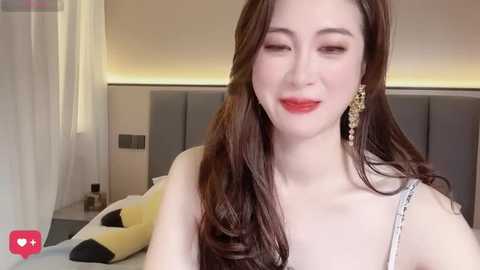 A video of a young woman with fair skin, long brown hair, and red lipstick, wearing a white sleeveless dress with silver embellishments, standing in a modern bedroom with a gray headboard.