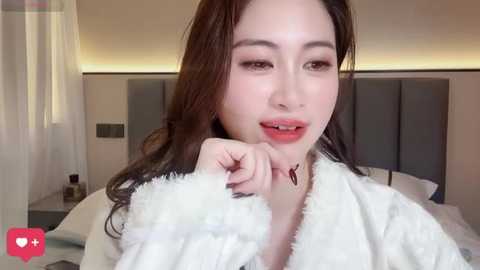 Video of an East Asian woman with long brown hair, fair skin, and red lipstick, wearing a white fluffy robe, in a modern bedroom with a gray upholstered headboard.
