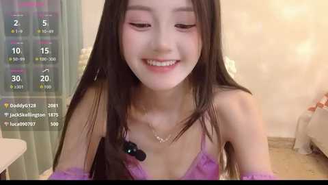 A young East Asian woman with long black hair and fair skin, wearing a purple lace bra, smiles in a cozy, dimly lit room. The image includes social media icons and a clock.